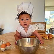 Idriss a rejoint le concours — aidez-le/la à gagner de superbes lots ! baby, child, chef_hat, paper_hat, flour, baking, kitchen, table, eggs, mixing_bowl, croissants, wooden_sign, curly_hair, white_shirt, indoor, person, face, wide_eyes, cooking, food