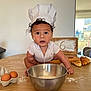 baby, child, chef_hat, paper_hat, flour, baking, kitchen, table, eggs, mixing_bowl, croissants, wooden_sign, curly_hair, white_shirt, indoor, person, face, wide_eyes, cooking, food