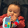 baby, child, toy, phone, red_sweater, wristband, colorful, indoor, closeup, face, hand, curly_hair, seat, playful, infant, cute, focused, soft_lighting, portrait, person