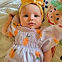 baby, child, headband, yellow, dress, orange_fruit, pattern, quilt, blanket, lying_down, cute, face, skin, infant, small_hand, colorful, soft_texture, indoors, portrait, young