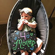 Nylah is registered to the contest to win money with this photo: baby, infant, headband, green_clothing, black_rocker, chalkboard_sign, indoor, carpet, baby_seat, bare_feet, cute, child, decorative_sign, colorful_text, hands_near_mouth, curious_expression, soft_lighting, floor, baby_accessory, sitting