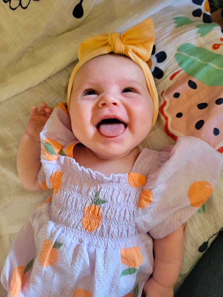 Nylah is registered to the contest to win money with this photo: baby, child, smiling, tongue_out, headband, yellow_headband, bow, dress, orange_pattern, fruit_pattern, lying_down, blanket, colorful_blanket, happy, cute, infant, pale_skin, short_hair, indoors, portrait