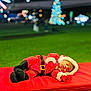 Carter is registered to the contest to win money with this photo: baby, sleeping, santa_outfit, christmas, holiday, infant, red_clothing, festive, outdoor, night, lights, blurred_background, green_grass, bench, cute, cozy, hat, gloves, face, child