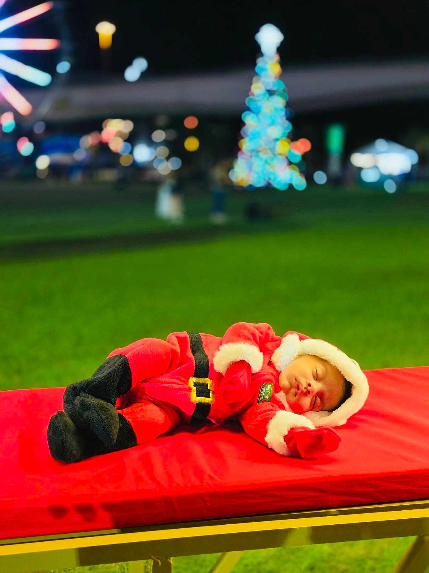 Carter is registered to the contest to win money with this photo: baby, sleeping, santa_outfit, christmas, holiday, infant, red_clothing, festive, outdoor, night, lights, blurred_background, green_grass, bench, cute, cozy, hat, gloves, face, child