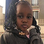 Marie participe au concours pour gagner de l'argent avec cette photo : child, pigtails, braided_hair, coat, outdoor, building, window, fence, face, portrait, playful, expression, young, person, clothing, street, daylight, cute, jacket, pout