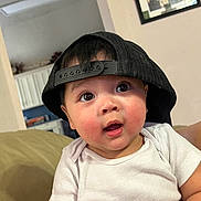 Lincoln joined the competition — help win amazing prizes! baby, child, cap, hat, indoor, couch, white_shirt, curious, wide_eyes, face, person, infant, skin, cheeks, short_sleeve, furniture, living_room, casual, portrait, cute