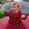 blond_hair, car, casual, child, cloudy_sky, cute, daytime, expression, face, green_object, hood, motion, outdoor, person, plaid, red_dress, suburban, toddler, trees, young