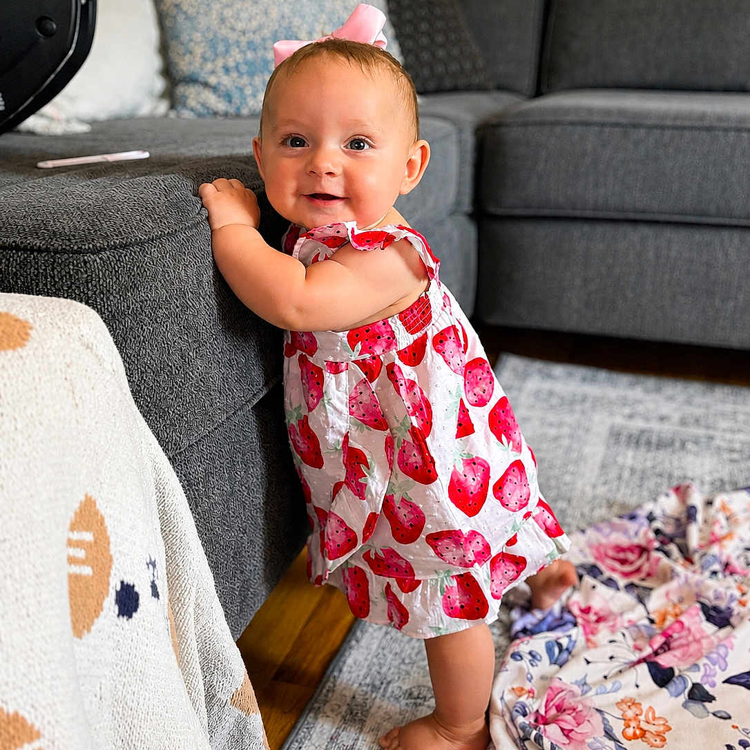 Ella-Grey joined the competition — help win amazing prizes! baby, child, girl, standing, smiling, pink_bow, strawberry_dress, couch, living_room, blanket, cushion, floor, carpet, wooden_floor, happy, indoor, portrait, furniture, home, cute