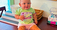 Ella-Grey is registered to the contest to win money with this photo: baby, child, pink_bow, green_shirt, new_balance, pink_shorts, table, wooden_table, basket, wall_art, red_truck, blessed_text, indoors, smiling, cute, sitting, person, face, happy, decor