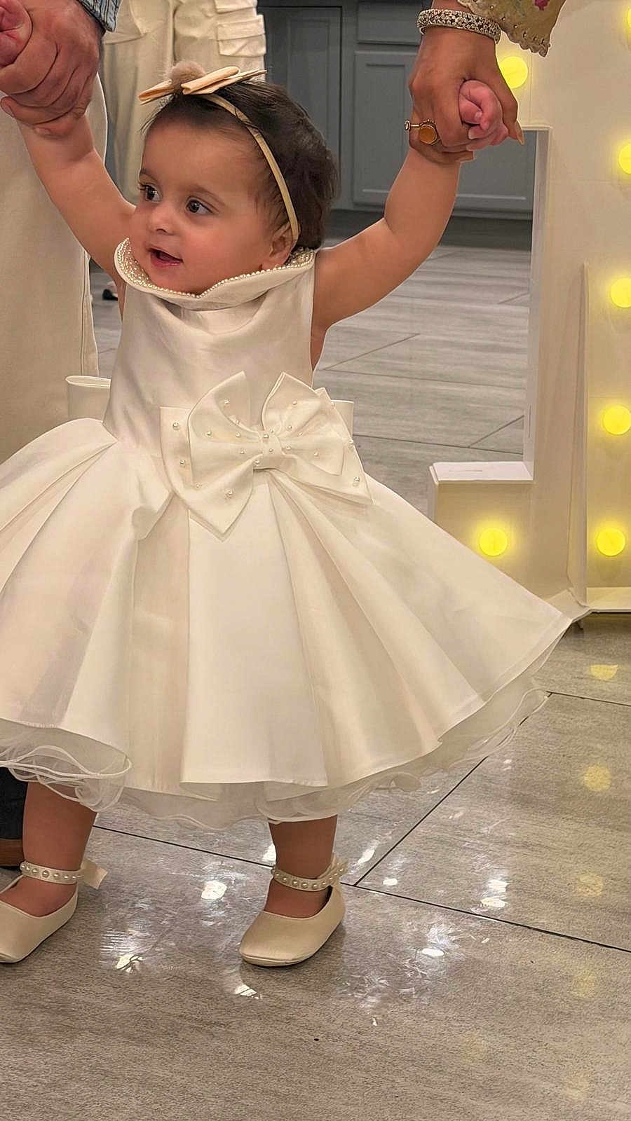 Sakina is registered to the contest to win money with this photo: toddler, child, white_dress, bow, headband, pearl, shoes, floor, tile, indoor, holding_hands, person, baby, celebration, party, cute, smiling, walking, fashion, accessories