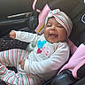 Delilah is registered to the contest to win money with this photo: baby, child, car_seat, smile, happy, headband, striped_clothing, infant, cute, portrait, indoors, seatbelt, clothing, person, car_interior, pink, white, striped_pants, young_child, playful