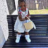 child, girl, standing, bench, white_dress, braided_hair, beads, handbag, sneakers, socks, outdoor, fence, window, wall, grass, sunlight, smile, portrait, accessories, fashion