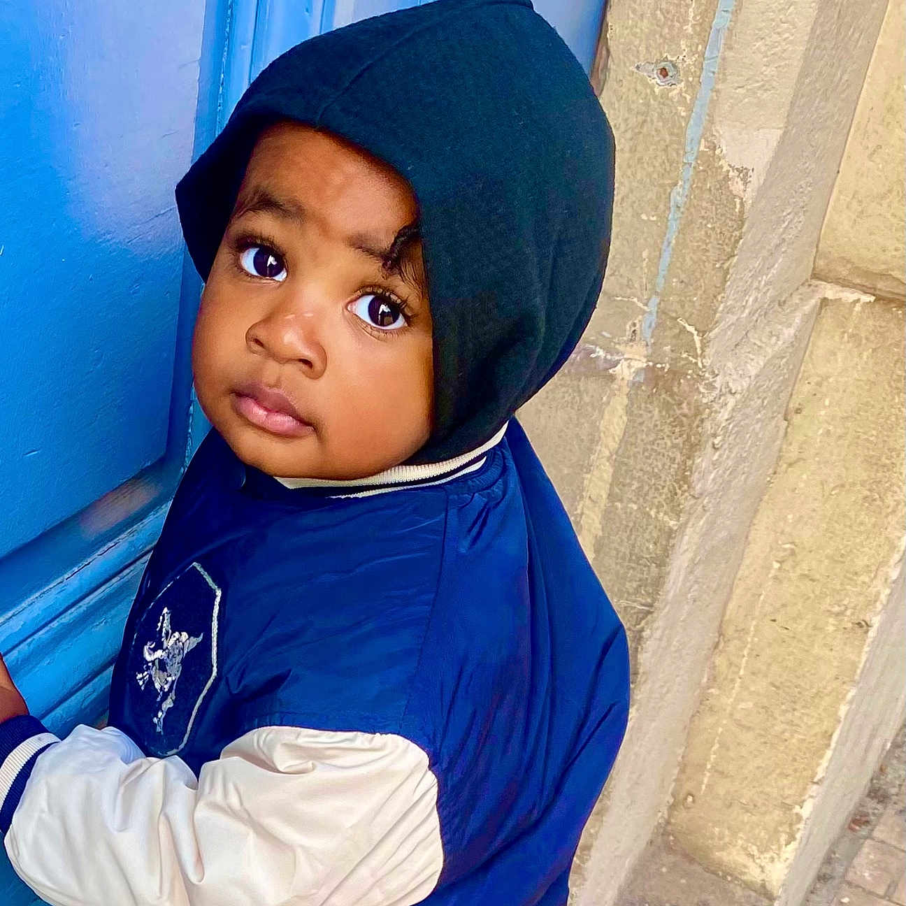 Kiene participe au concours pour gagner de l'argent avec cette photo : blue_door, building, child, coat, curious_gaze, face, hand_on_door, hood, hooded_jacket, jacket, jeans, outdoor, person, portrait, standing, stone_wall, street, toddler, urban, wide_eyes