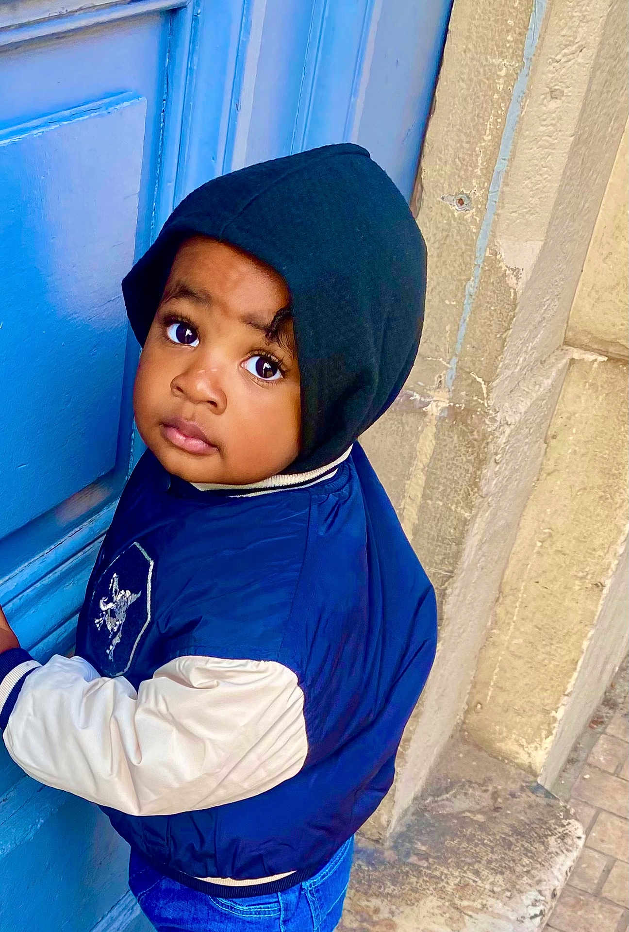 Kiene participe au concours pour gagner de l'argent avec cette photo : child, toddler, hood, hooded_jacket, jacket, blue_door, curious_gaze, wide_eyes, stone_wall, jeans, standing, portrait, outdoor, hand_on_door, urban, person, face, coat, street, building