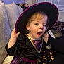 Connie is registered to the contest to win money with this photo: child, costume, witch_hat, purple_dress, stars, gold_trim, couch, indoor, curtains, curly_hair, expression, sitting, face, person, home, cozy, living_room, cute, holiday, celebration