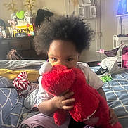 Raya is registered to the contest to win money with this photo: child, plush_toy, elmo, bed, blanket, indoor, hair, clothing, person, toy, furniture, room, sitting, hand, face, footwear, decor, television, door, cushion