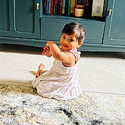 Adya is registered to the contest to win money with this photo: toddler, child, white_dress, carpet, rug, indoor, cabinet, books, furniture, curly_hair, person, carpet_pattern, floor, sitting, looking_back, home, cozy, cute, young_child, living_room