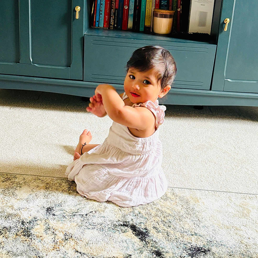 Adya is registered to the contest to win money with this photo: books, cabinet, carpet, carpet_pattern, child, cozy, curly_hair, cute, floor, furniture, home, indoor, living_room, looking_back, person, rug, sitting, toddler, white_dress, young_child