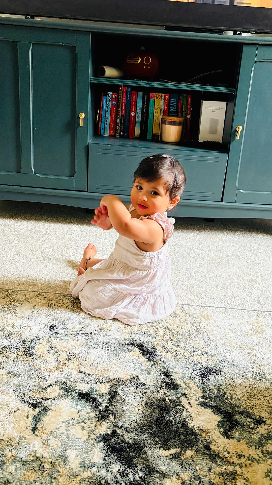 Adya is registered to the contest to win money with this photo: toddler, child, white_dress, carpet, rug, indoor, cabinet, books, furniture, curly_hair, person, carpet_pattern, floor, sitting, looking_back, home, cozy, cute, young_child, living_room