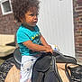 child, toddler, curly_hair, blue_shirt, panda_pants, saddle, outdoor, sunlight, brick_wall, white_door, bench, footwear, focused, cute, young_child, playful, casual_clothing, daylight, person, portrait