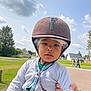 child, helmet, cardigan, outdoor, sky, cloud, grass, person, hand, walking, path, houses, daytime, casual_clothing, greenery, support, curious_expression, young_child, nature, sunny