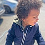 child, toddler, curly_hair, smiling, jacket, shopping_cart, outdoor, daylight, happy, person, clothing, pavement, car, blurred_background, portrait, cute, young_child, casual_wear, joyful, fun