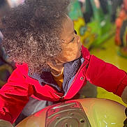 Louka a rejoint le concours — aidez-le/la à gagner de superbes lots ! child, curly_hair, red_jacket, ride, amusement_park, colorful, indoor, play, portrait, side_profile, young, fun, vibrant, clothing, person, activity, focus, light, blurred_background, casual
