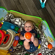 Messiah is registered to the contest to win money with this photo: baby, infant, child, playpen, ball, orange_ball, toys, blanket, plush_blanket, indoor, portrait, sitting, wide_eyes, looking_at_camera, blue_clothing, colorful, playmat, animal_illustration, wood_floor, happy