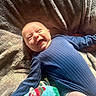 baby, smiling, blue_outfit, blanket, cozy, texture, soft, sunlight, happy, infant, lying_down, cute, child, indoors, warm_light, blanket_pattern, comfort, portrait, playful, relaxed