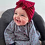 baby, child, smiling, red_headwrap, bow, patterned_clothing, seated, cushion, indoor, happy, cute, portrait, infant, chubby_cheeks, long_sleeves, buttoned_shirt, bright, warm, closeup, face