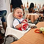 baby, high_chair, smiling, child, outdoor, dining, table, toy, person, happy, plastic_bag, short_sleeve_shirt, people, restaurant, summer, daylight, casual, food, seat, background