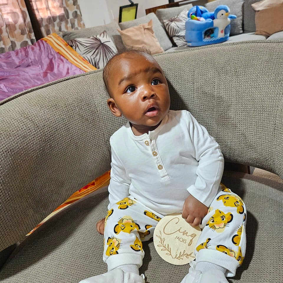 Hysaël Kenal participe au concours pour gagner de l'argent avec cette photo : baby, child, couch, cozy, curtains, cute, expression, furniture, home, indoors, infant, milestone, patterned_pants, penguin_toy, pillow, plush_toy, seated, sofa, white_shirt, wooden_plaque