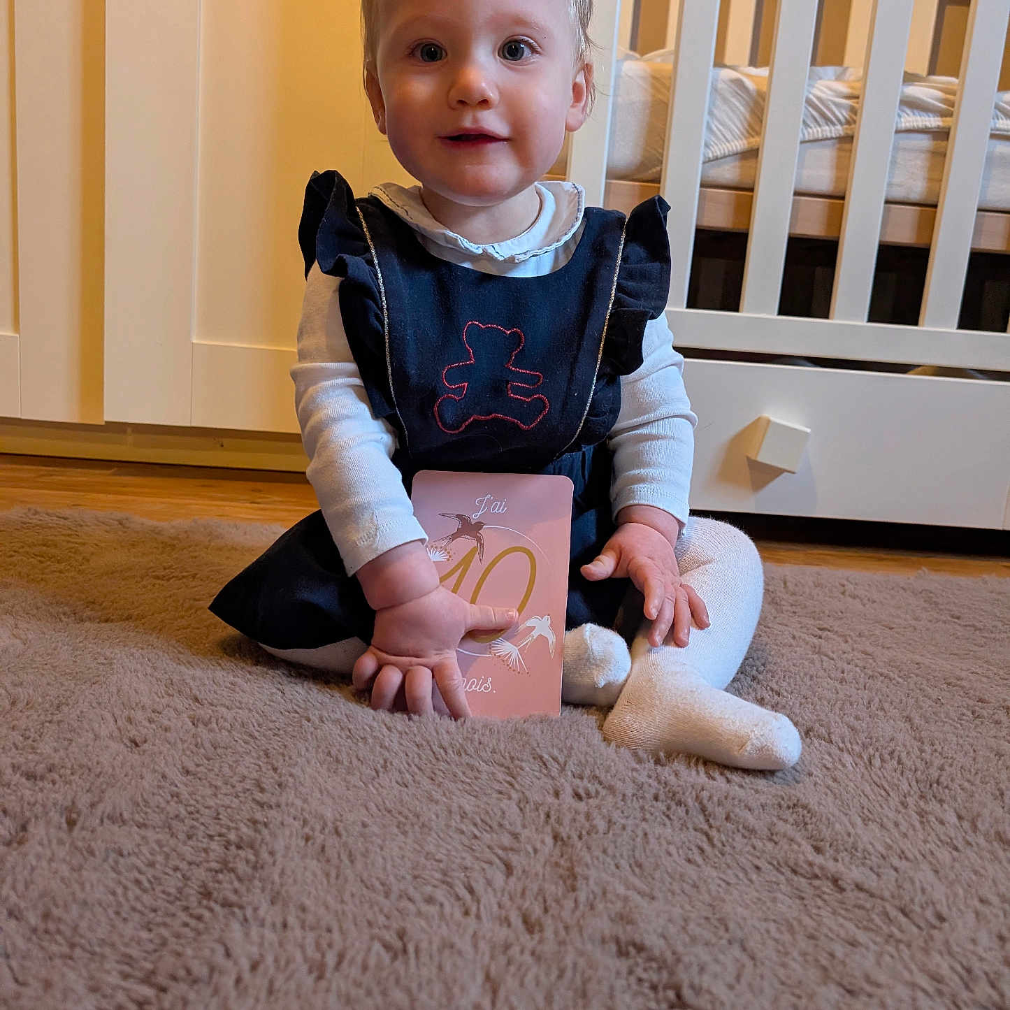 Maryléa a rejoint le concours — aidez-le/la à gagner de superbes lots ! baby, child, toddler, carpet, dress, white_tights, crib, room, floor, furniture, card, person, hair_tie, indoor, cute, smiling, sitting, toy, wooden_floor, soft_lighting