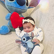 Olivia participe au concours pour gagner de l'argent avec cette photo : baby, blanket, child, couch, cozy, cute, flower_pattern, headband, heart, indoor, infant, plush_toy, red_heart, seated, smile, soft_texture, stitch, stuffed_animal, toy, white_outfit