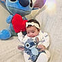 baby, blanket, child, couch, cozy, cute, flower_pattern, headband, heart, indoor, infant, plush_toy, red_heart, seated, smile, soft_texture, stitch, stuffed_animal, toy, white_outfit