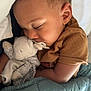 child, toddler, sleeping, blanket, pacifier, stuffed_animal, bed, cozy, cute, face, curly_hair, brown_shirt, indoor, rest, comfort, nap, soft_toy, peaceful, closeup, relaxation