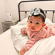 Jannatul is registered to the contest to win money with this photo: baby, infant, pink_clothing, headband, silver_bow, bed, white_bedding, pillow, indoors, furniture, nightstand, curious_expression, person, child, cute, portrait, sleepwear, headwear, lying_down, room