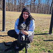 Pippin is registered to the contest to win money with this photo: animal, beanie, black_dog, casual_clothing, crouching, daylight, dog, fence, friendship, grass, happy, leisure, nature, outdoor, pet, smiling, sunny, trees, winter, woman