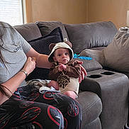 Desmond is registered to the contest to win money with this photo: adult_hand, baby, baby_toy, carpet, casual, clothing, couch, curtains, face, furniture, hoodie, indoor, infant, living_room, pajamas, person, picture_frame, sitting, toy, window