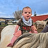 animal, baby, bib, camo_clothing, child, cloud, drool, farm, fence, hand, hoofed_animal, horse, outdoor, person, pet, ranch, rural, sky, tree, vehicle