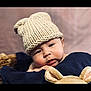 baby, child, knitted_hat, plush_toy, cushion, face, chubby_cheeks, indoor, portrait, cute, soft_toy, headwear, resting, baby_clothing, person, closeup, warm_colors, cozy, infant, expression