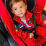 child, toddler, car_seat, red_clothing, seatbelt, car_interior, looking_away, racing_patch, young_child, sitting, safety_harness, cute, portrait, casual, daylight, vehicle, young_boy, curious_expression, indoors, seat