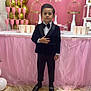 child, boy, tuxedo, bow_tie, party, birthday, celebration, pink, balloons, champagne_glasses, table, decorations, formal_wear, marble_floor, standing, portrait, event, young, indoors, smile
