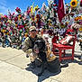 soldier, dog, service_dog, camouflage, vest, flowers, memorial, flag, red_chair, stuffed_animals, sidewalk, sunny_day, blue_sky, outdoor, person, pet, kneeling, sunglasses, concrete, nature