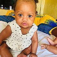 Ava joined the competition — help win amazing prizes! baby, child, infant, sitting, wide_eyes, hair_puffs, white_dress, polka_dots, bed, pillows, yellow_pillow, blue_pillow, skin, cute, indoors, furniture, room, foot, hand, expression