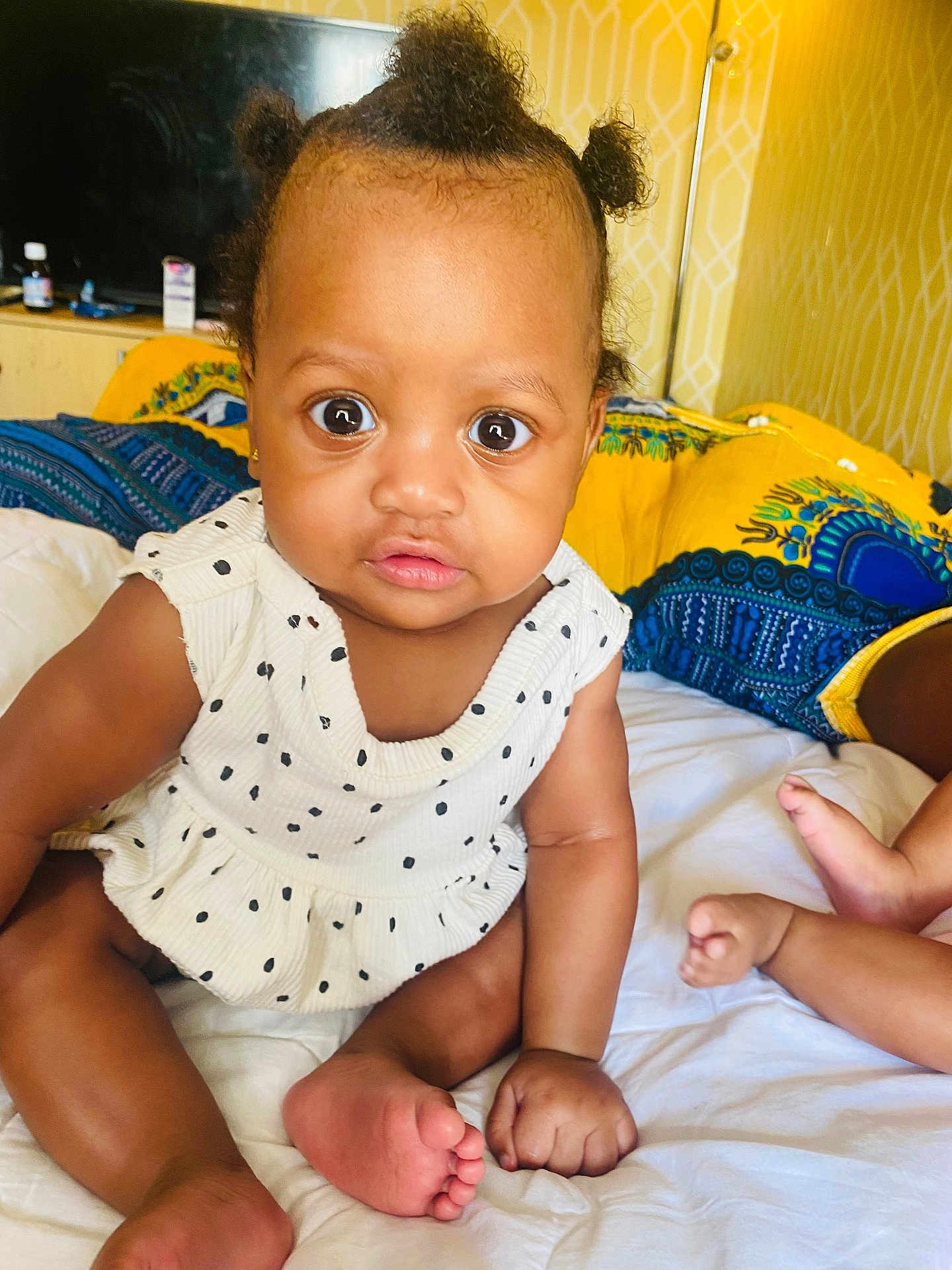 Ava joined the competition — help win amazing prizes! baby, child, infant, sitting, wide_eyes, hair_puffs, white_dress, polka_dots, bed, pillows, yellow_pillow, blue_pillow, skin, cute, indoors, furniture, room, foot, hand, expression