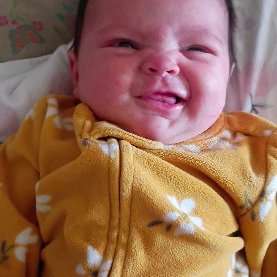 Hazel-Lee joined the competition — help win amazing prizes! baby, infant, onesie, yellow_clothing, floral_pattern, bed, blanket, cute, face, tongue_out, scrunched_eyes, child, soft_lighting, indoors, portrait, expression, lying_down, young_child, funny_face, cozy