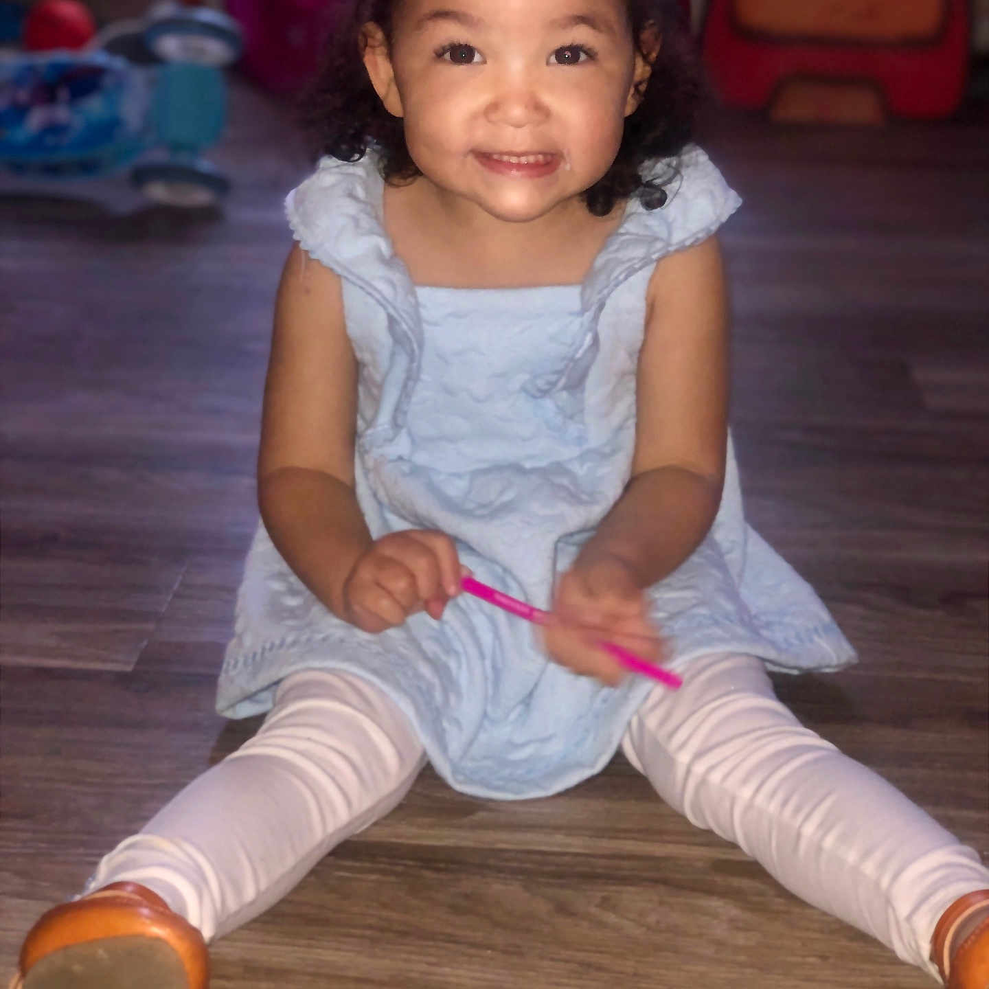 Kamara joined the competition — help win amazing prizes! blue_dress, brown_shoes, child, curly_hair, cute, face, girl, happy, holding, indoor, person, pink_object, playful, portrait, sitting, smiling, toy, white_leggings, wooden_floor, young