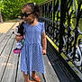 child, girl, sunglasses, blue_dress, plush_toy, minnie_mouse, wooden_bridge, black_iron_railings, greenery, outdoor, summer, smiling, curly_hair, orange_shoes, walking, nature, sunlight, cute, person, portrait