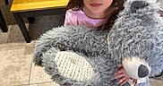 Callie is registered to the contest to win money with this photo: child, teddy_bear, toy, indoor, kitchen, table, bench, tile_floor, curly_hair, pink_shirt, stuffed_animal, person, looking_up, plush, home, furniture, carpet, cute, young, holding